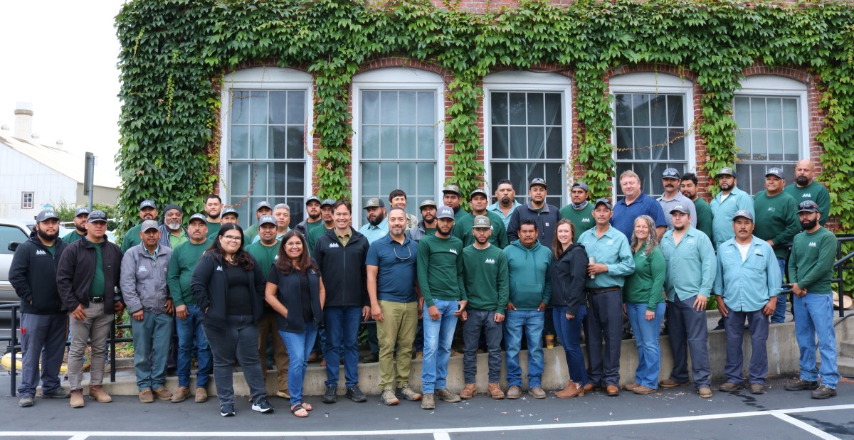 Penning Company Photo (Cropped) Penning Landscapes team in green workwear posing in front of a historic brick building with climbing ivy.